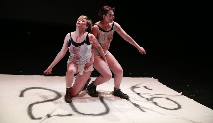 Two white women standing on a white sheet that has "2016" written on it. The two women are falling down with blood on them.