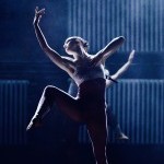 A dancer stands on a dimly lit stage, illuminated by cool blue lighting. She balances on one leg while lifting the other.