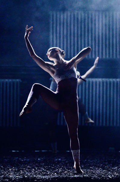 A dancer stands on a dimly lit stage, illuminated by cool blue lighting. She balances on one leg while lifting the other.