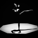 A dancer stands on a spotlighted stage, surrounded by darkness. One leg is extended horizontally behind them, while their arms a