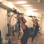 Group class all standing with arms stretching overhead with arms in straps. 