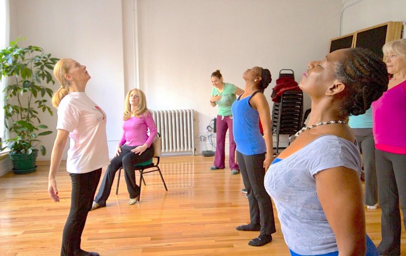 Dancers facing instructor, lifting their chests to the sky with arms by their side, all standing but one in a pink shirt sitting