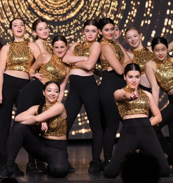 Group of young dancers posed around each other with gold sequin top and black bottoms. 