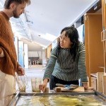 Woman speaking to a young man about artwork on a table.