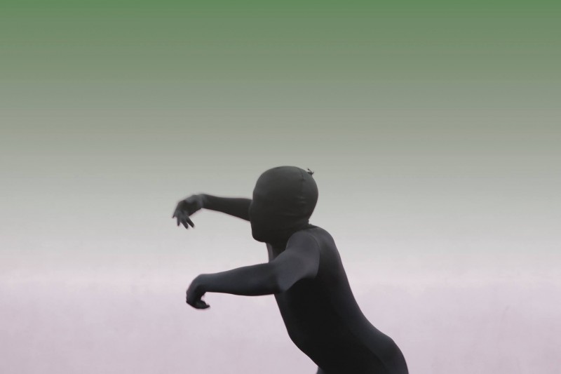 Profile of person in a black skintight body suit lifting their arms. There is a pink and green washing in the background.