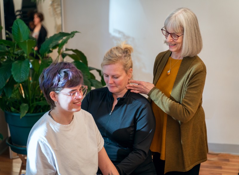 Three people, two seated & one standing behind, are shown smiling during an Alexander Technique class at the Balance Arts Center