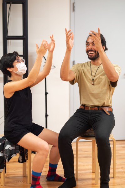 two students sit on stools, smiling, with hands in the air 