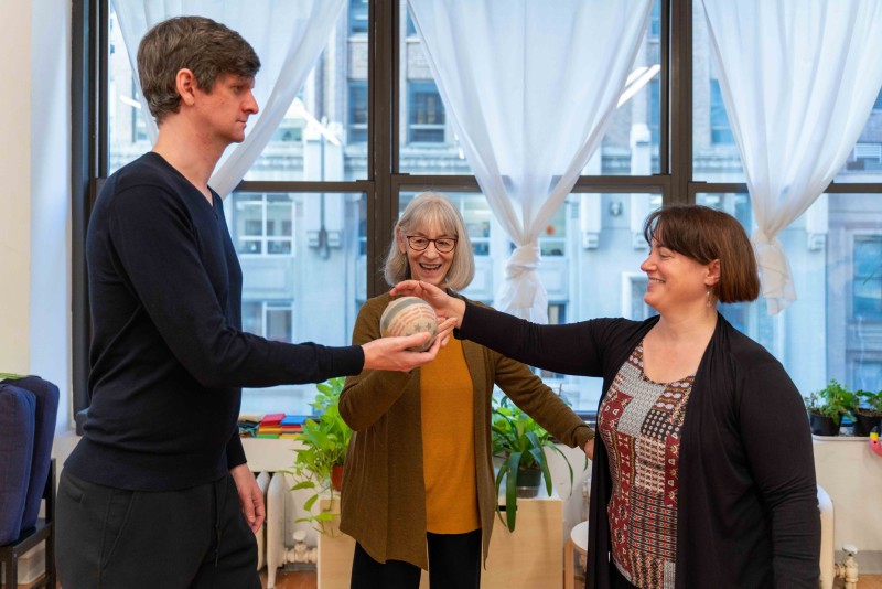 A smiling Alexander Technique teacher guiding two teacher trainees to pass a ball back and forth with good use and awareness. 