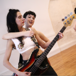 Dancer hugs base player from behind while singing and slyly eyeing the camera.