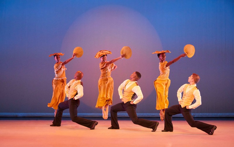 Four female dancers in yellow dresses hold fans and jump while four male dancers kneel in front of them