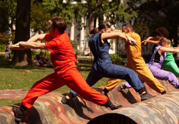 Three brightly dressed dancers on top of an outdoor art piece, stretching over the waved steel structure, resembling a carpet