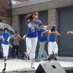Singer holding a microphone with dancers in the background all wearing blue tops and white pants