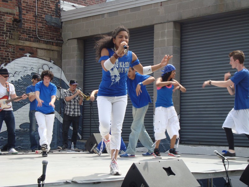 Singer holding a microphone with dancers in the background all wearing blue tops and white pants