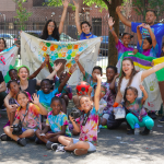 A small crowd of children and adults are gathered, smiling in colorful clothing with banners that say "Dream, Create, Thrive"