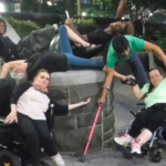 Two white women in wheelchairs leaning toward a stone wall. Two black men, an Asian man and white elder woman draped atop it.