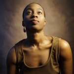 African american woman with very short hair and brown dress looks upward as she poses for a portrait against an brown backdrop.