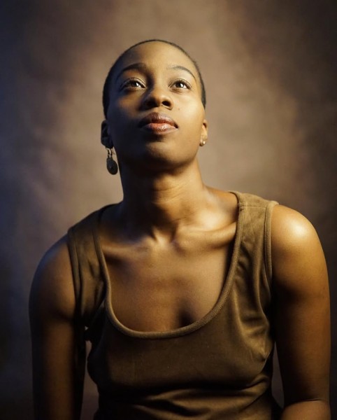 African american woman with very short hair and brown dress looks upward as she poses for a portrait against an brown backdrop.