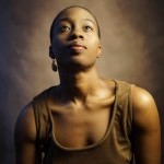 Black woman with short hair in brown dress poses in front of brown back drop looking up in a portrait photo.