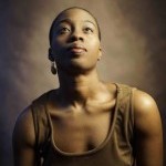 Black woman with short hair in brown dress poses in front of brown back drop looking up in a portrait photo.