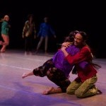 image of 2 performers embracing and smiling on the ground in bright, colorful clothing