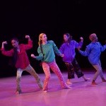 4 dancers on stage in colorful clothing, elbows bent in a variety of muscle poses, expressions of care in their eyes.