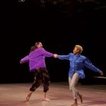 Two dancers in loose, colorful shirts hold hands and swing in opposing circles, smiling playfully at each other.