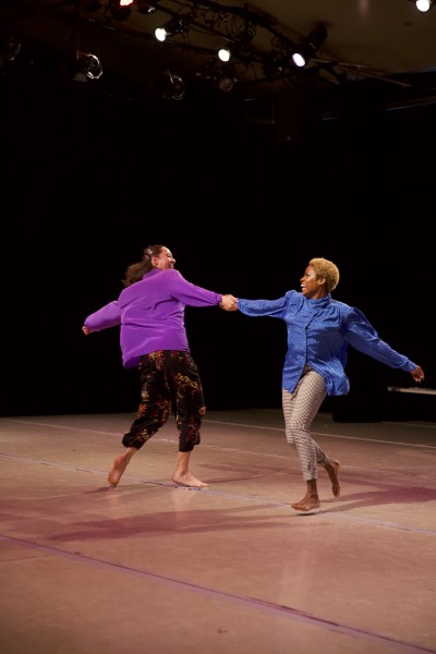 Two dancers in loose, colorful shirts hold hands and swing in opposing circles, smiling playfully at each other.