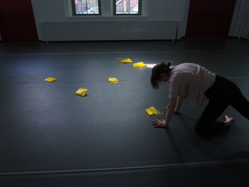 Female dancer on hands and knees, surrounded by squares of folded thin yellow plastic, in a studio with light from windows 