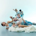 Dancers tumbling to the floor in a coiled group, wearing blues and whites, eyes closed, immersed in the sensation of a wave