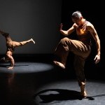 Two dancers in separate spotlights. Dancer on the left is in a hand stand, dancer on the right is standing with one leg up.