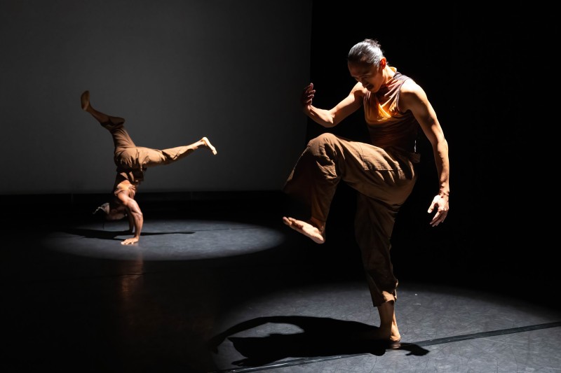 Two dancers in separate spotlights. Dancer on the left is in a hand stand, dancer on the right is standing with one leg up.