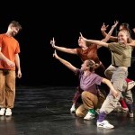 4 dancers huddle in a group, gesturing hand towards another dancers smiling towards them