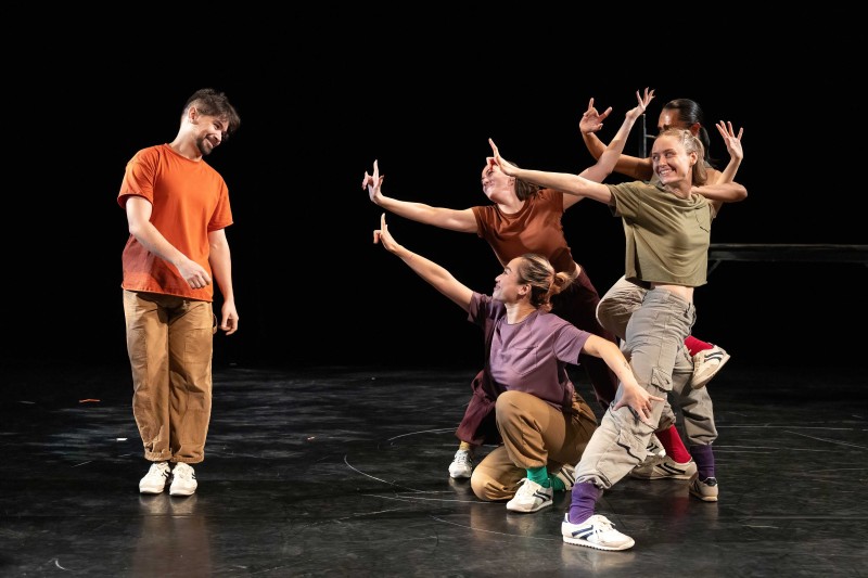 4 dancers huddle in a group, gesturing hand towards another dancers smiling towards them