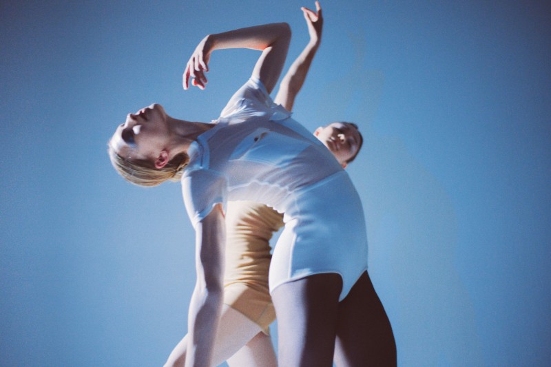 Two dancers in white and pale yellow pose in fluid, arching poses against a soft blue backdrop, captured from a low angle