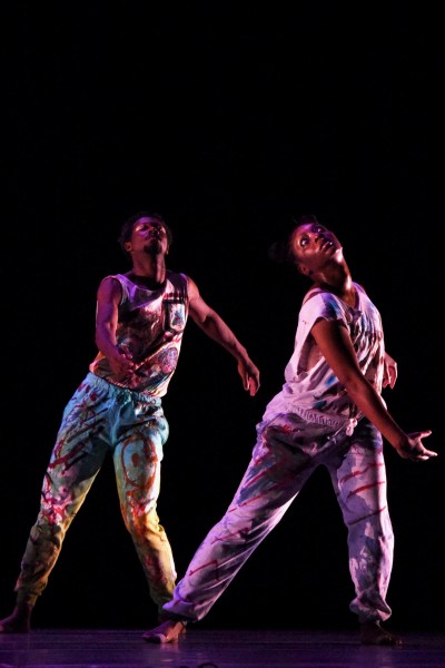 2 Black performers in multicolored sweats lean towards their left surrounded by darkness