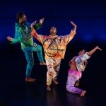 Three black performers in vibrant sweatsuits frame each other and refer to the stars