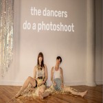 2 people sitting against a white wall with the text "the dancers do a photoshoot" above their heads.