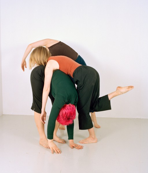 Three dancers fold on top of each other. One dancer has bright pink hair
