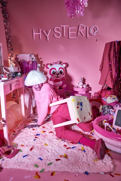 Raja Feather Kelly is sprawled on the floor of a messy pink room wearing a pink sweater, pink pants, and a white bowl cut wig