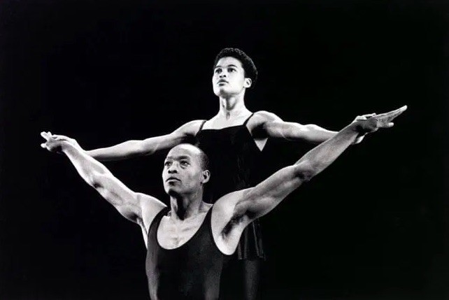 A black and white photo of Bill T. Jones crouching in front of Roz LeBlanc who is holding his wrists and spreading his arms wide