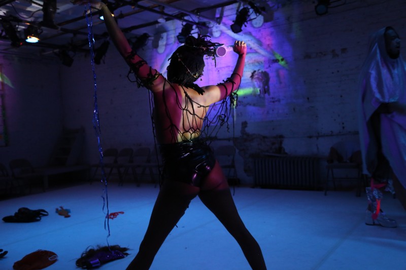 A masked performer draped in fabric reaches across the space. The back wall is lit with multicolor lights.
