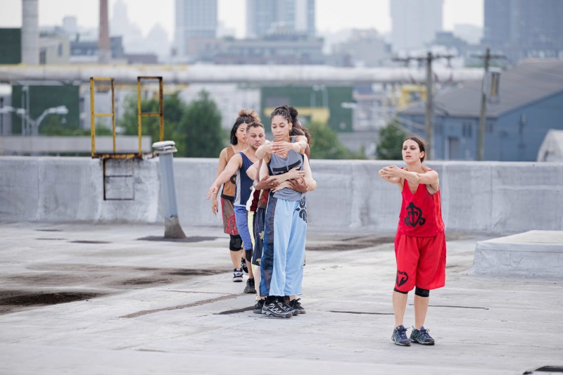Eight dancers in a line on a rooftop, each hugging the person in front of them, with the first dancer in red and second in blue 