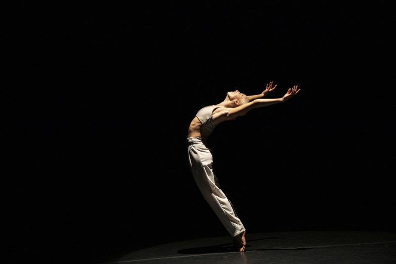 Dancer Zoey Anderson in a small backbend with both hands extended overhead while on a dark lit stage