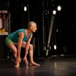 Photo of K.J. Holmes in a blue shirt and green shorts, in a deep plie hinging forward looking out towards a black stage