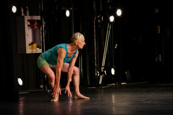 Photo of K.J. Holmes in a blue shirt and green shorts, in a deep plie hinging forward looking out towards a black stage