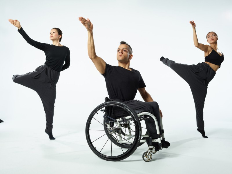 a photo of 3 dancers, one in a wheelchair, 2 jumping in the air. All 3 dancers have 1 arm in the air