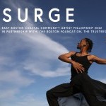 A male dancer, wearing a black shirt and pants, holds his right hand at his stomach. His left arms bends and lifts behind him.