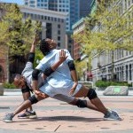 Male dancer 1 stands in a wide lunge position. Male dancer 2 intertwines his arms with dancer 1, also in a lunge.