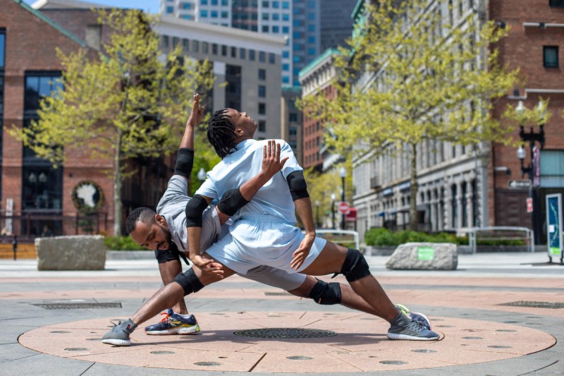 Male dancer 1 stands in a wide lunge position. Male dancer 2 intertwines his arms with dancer 1, also in a lunge.