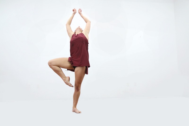 Woman in a short red dress standing on left leg with right leg in pase. Torso is arched back and arms are lifted up over head an